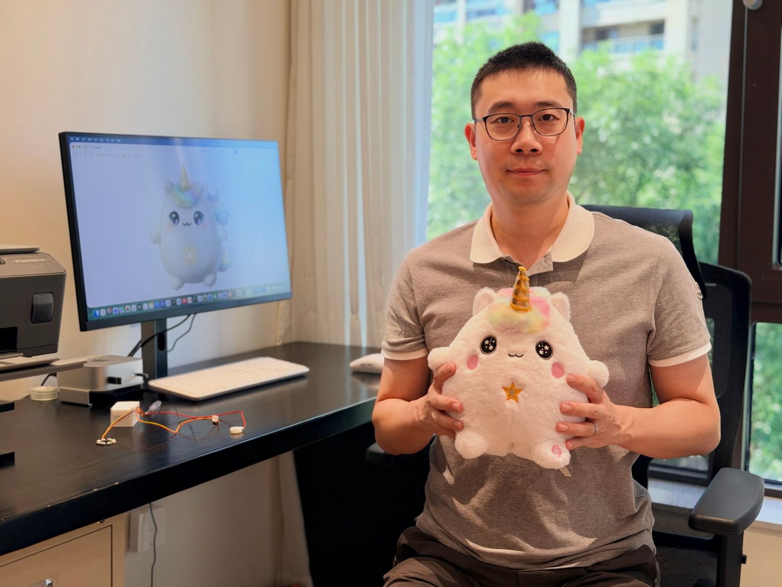 Yirun Chen at his desk holding Lumi, with the Lumi concept art on the monitor behind him
